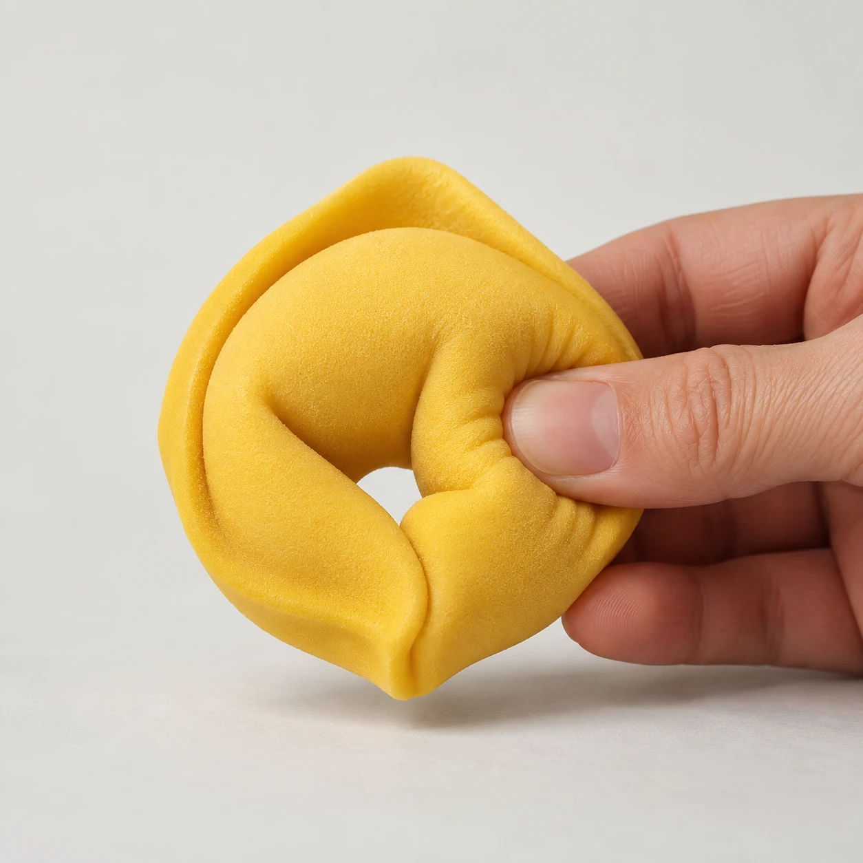 Studio photograph of a yellow foam tortellino stress toy visibly compressed by a hand.