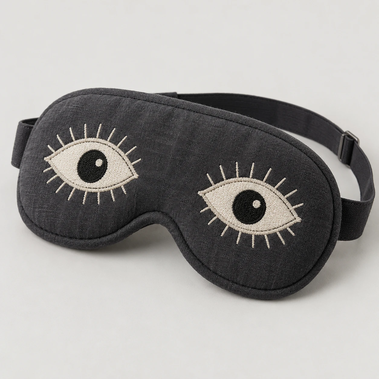 Charcoal padded sleep mask with two embroidered open eyes and an elastic strap.