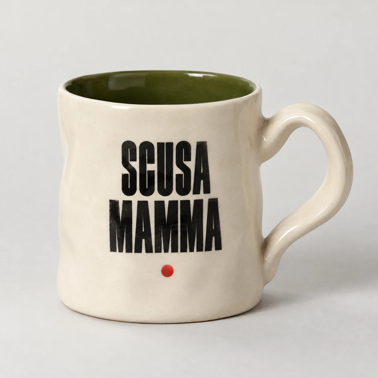 Studio photograph of an ivory ceramic mug with a curved handle, olive-green interior and black Scusa Mamma lettering.